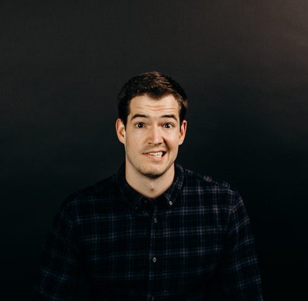 Comedian Andrew Stanley stands in a plaid shirt in front of a dark background with a smile/grimace.