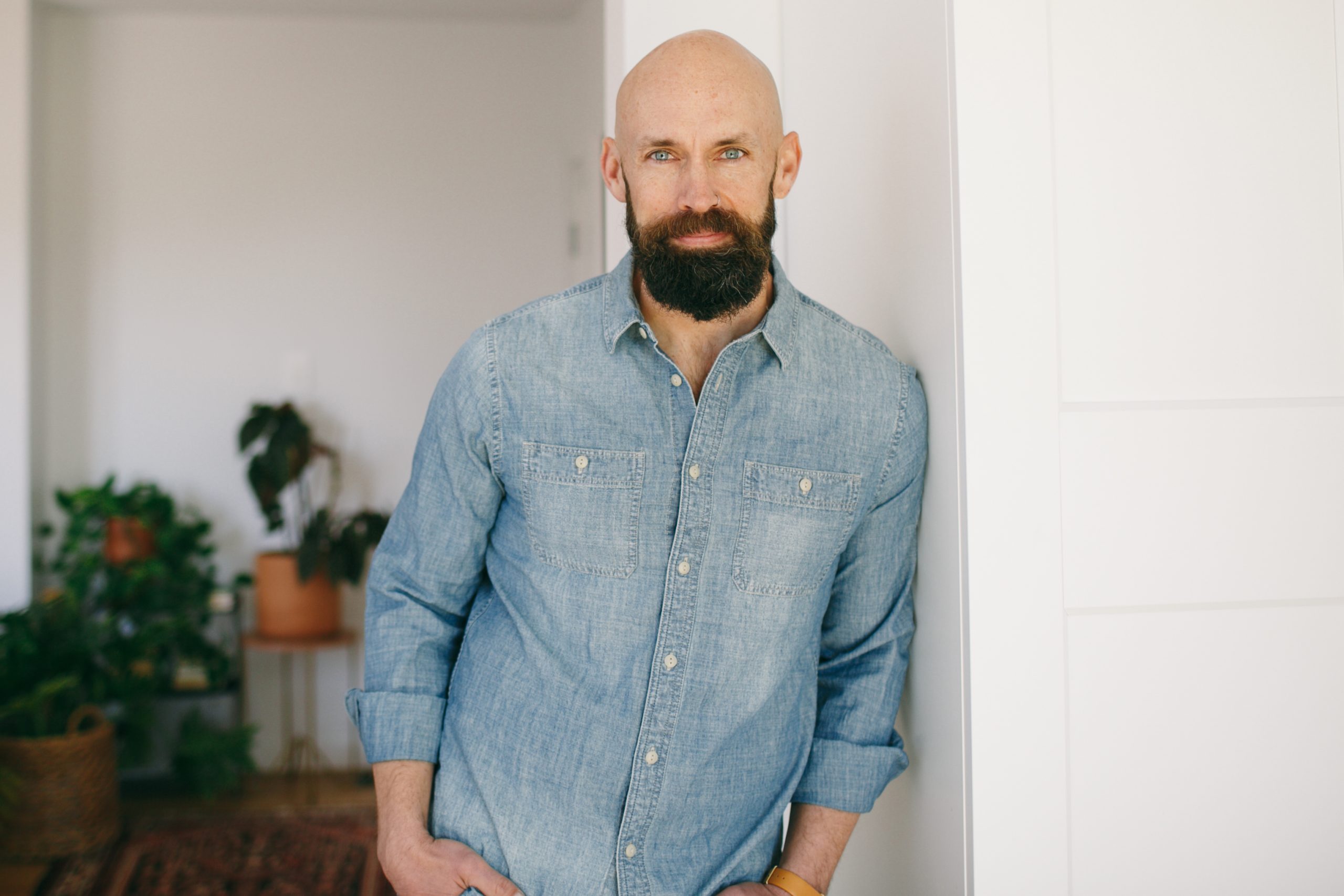 Joseph Barkley leans against a white wall in a denim shirt. Green plants are on a plant stand in the background.