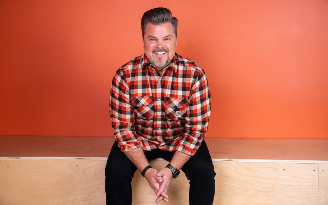 Tim Lucas sits smiling on a wooden surface in front of a bright orange background.
