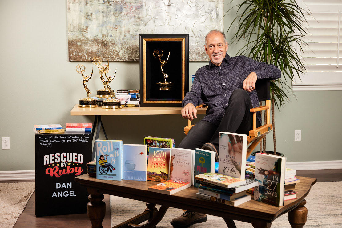 Dan angel sits in a director's chair with multiple awards and images of his work surrounding him.